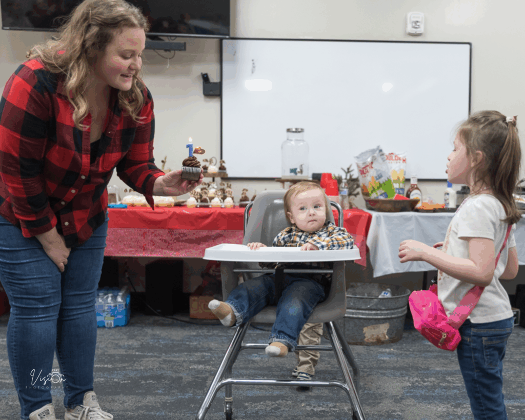 Image of mom holding cupcake while singing. Older sister ready to blow out candles.