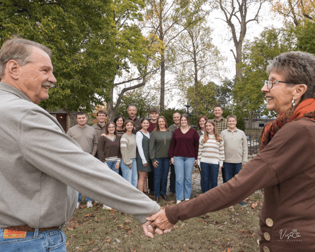 Image of husband and wife holding hands looking at one another while kids and grandkids look at camera